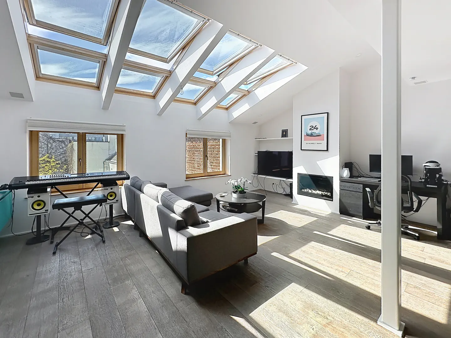 Bright, modern living room with skylights, gray sofa, keyboard, and desk areas. Hardwood floors and white walls create a spacious feel.
