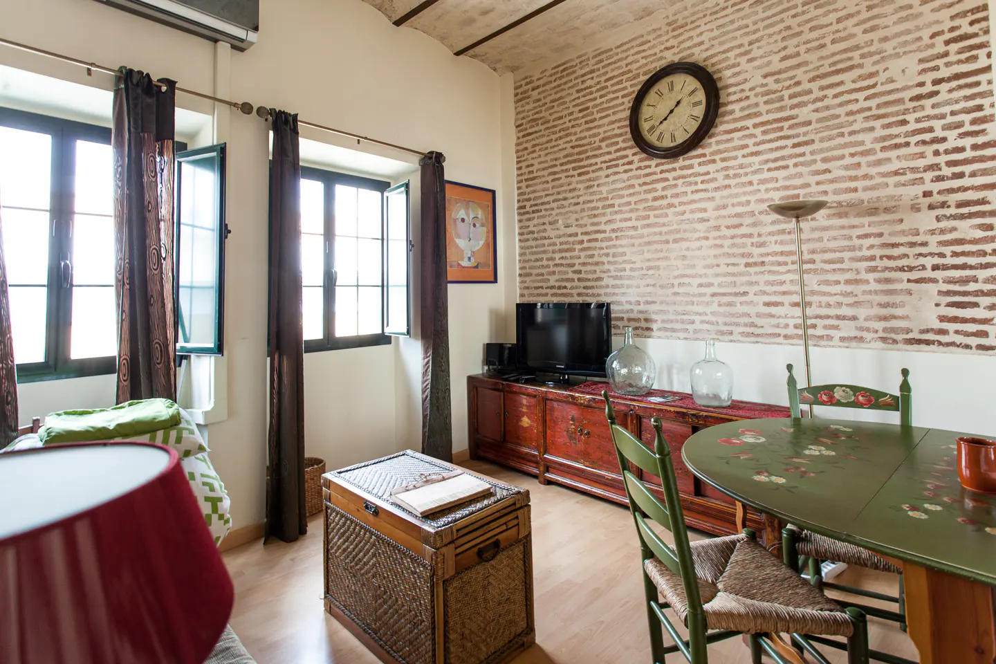 Living room with brick wall, wooden floors, and windows with brown curtains. A green table and chairs are on the right.