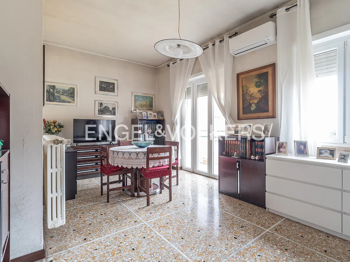 A bright living room with a round table, red chairs, and a terrazzo floor. Paintings and a TV are visible.