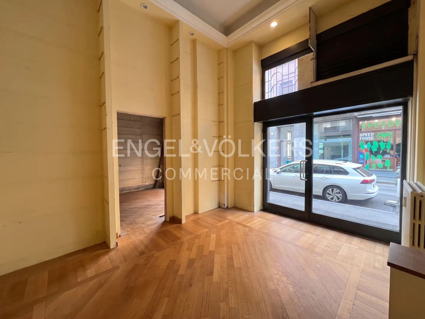 Empty commercial space with herringbone wood floors, yellow walls, and a glass storefront. A doorway leads to another room. A white car is visible outside.