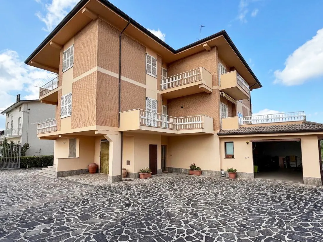 A three-story brick house with balconies and a stone driveway on a sunny day.