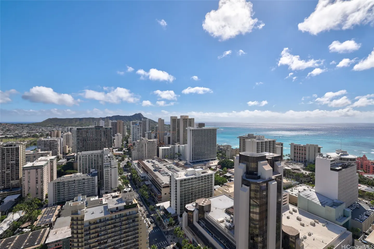 Aerial view of Honolulu, Hawaii, featuring Diamond Head, tall buildings, and the ocean under a blue sky with scattered clouds.
