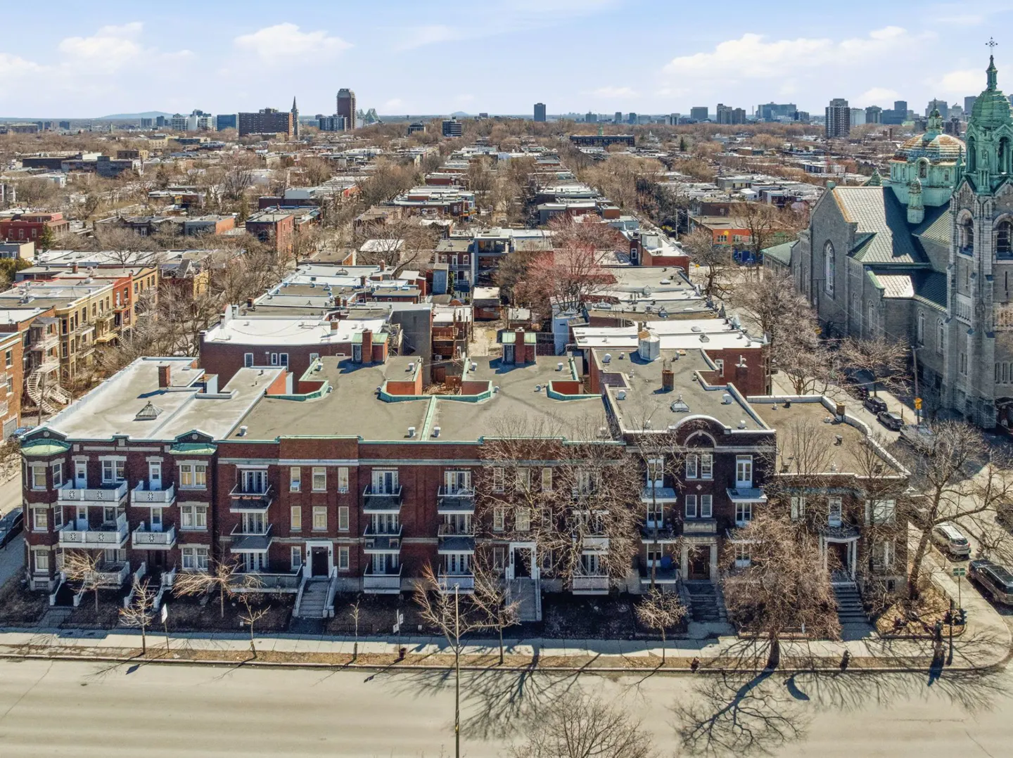 Aerial view of a red brick apartment building with balconies, a church, and a city skyline in the background.