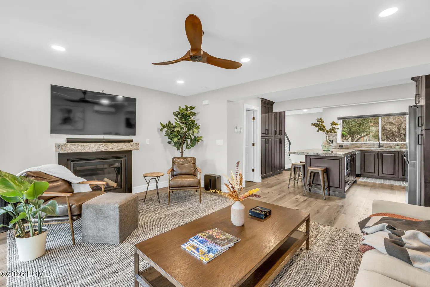 Open concept living room and kitchen with brown leather chairs, fireplace, and wood floors.