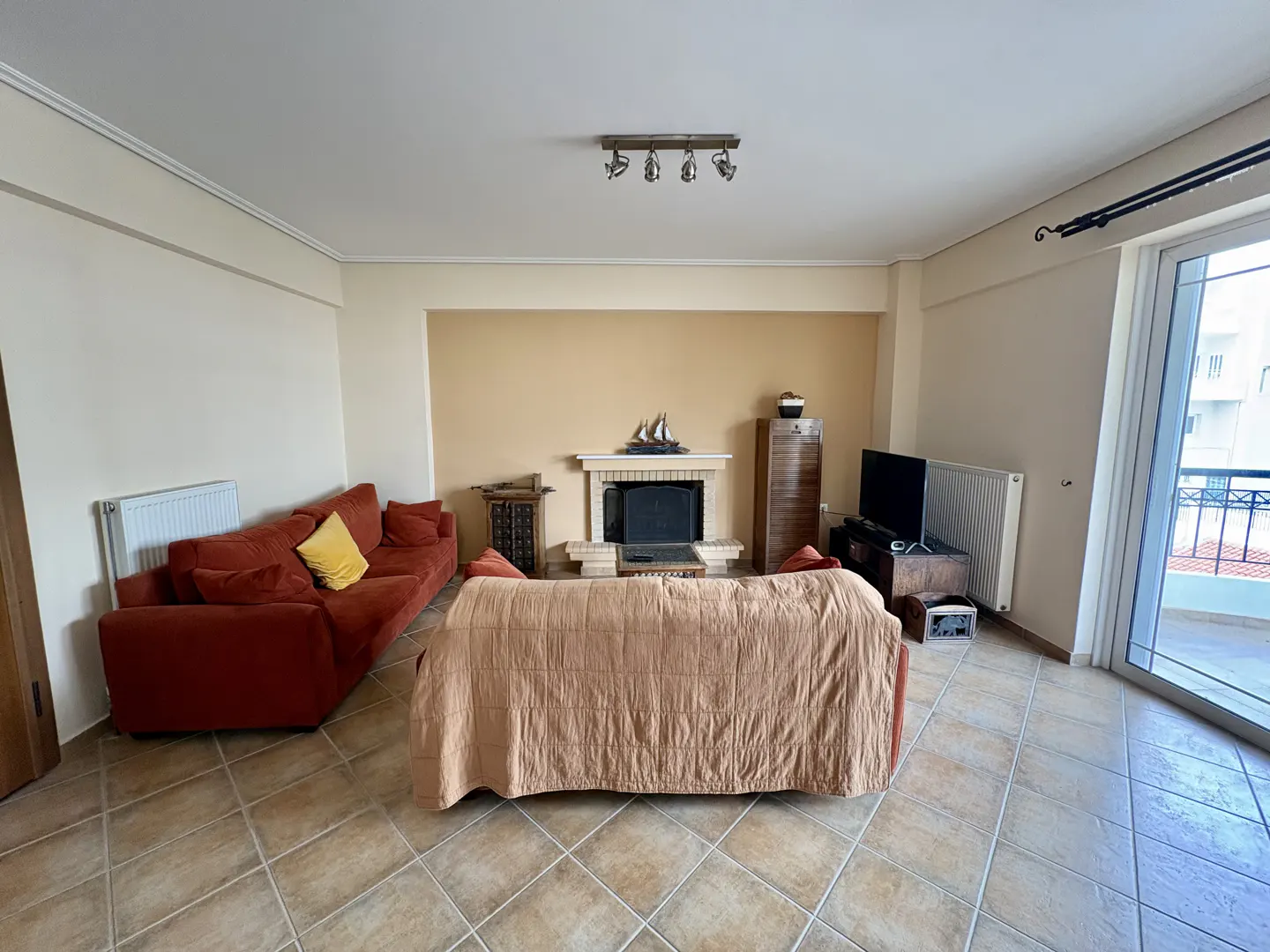 A living room with a fireplace, a red sofa with a yellow pillow, and a covered sofa. There is a TV and a balcony door.