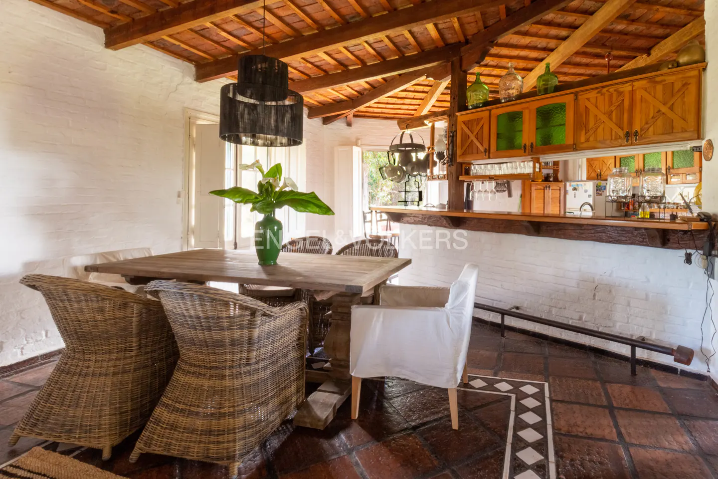 Rustic dining room with a wooden table, wicker chairs, and a bar area with wooden cabinets and glassware. The floor is tiled in terracotta.
