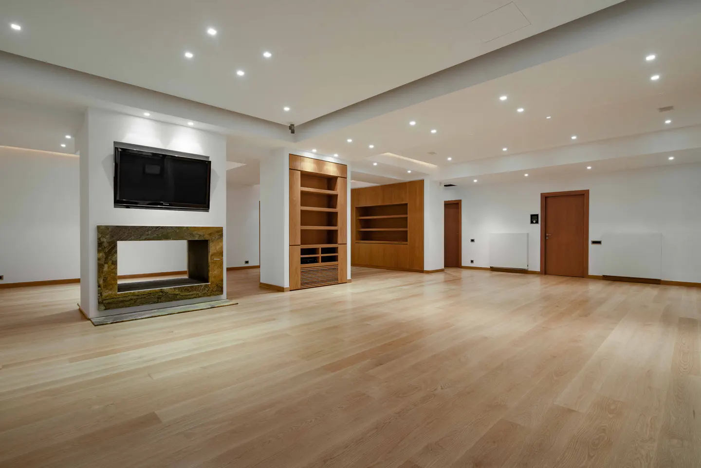 Bright, empty living room with light wood floors, white walls, recessed lighting, TV over stone fireplace, and built-in wooden shelving.
