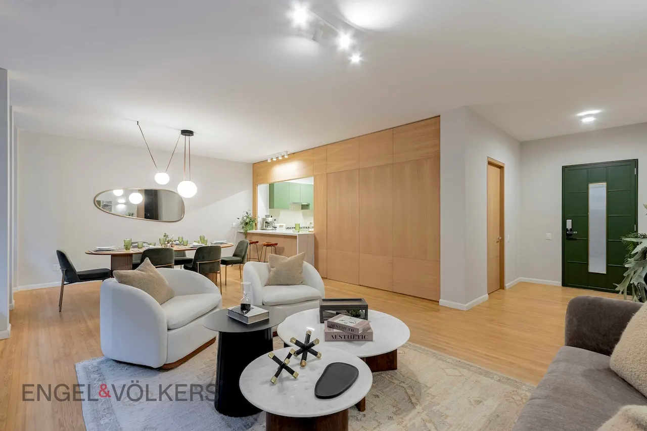 Open-concept living space with wood floors, white chairs, and a dining table. A green front door and kitchen are visible in the background.