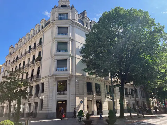 Exterior view of a light-colored, multi-story building with balconies and a Massimo Dutti store on the ground floor, framed by green trees.