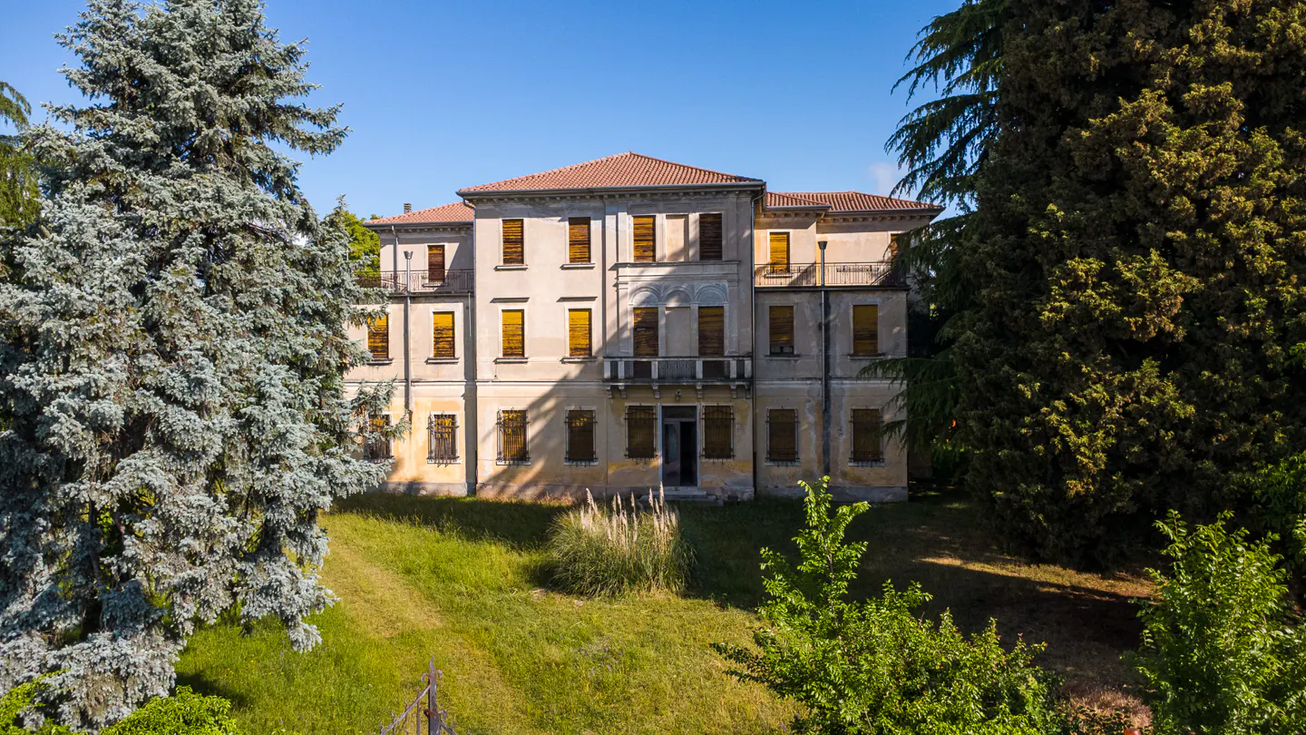 Exterior view of a three-story beige building with a red tile roof and yellow shutters, surrounded by green trees and grass.