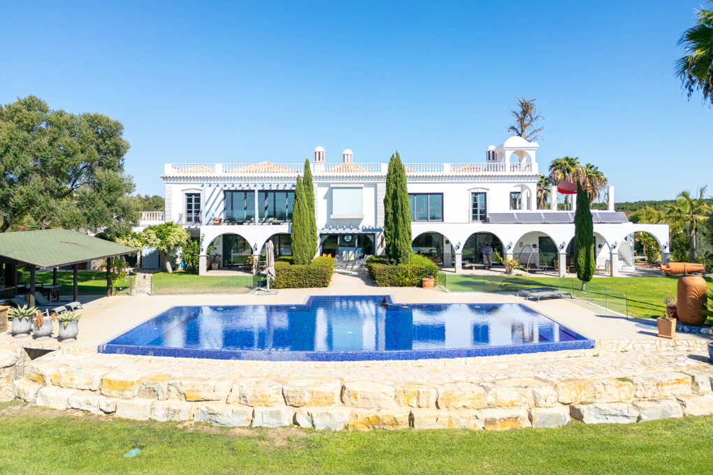 Large white villa with a blue swimming pool, stone patio, and lush green lawn under a clear blue sky. [AI-generated text]