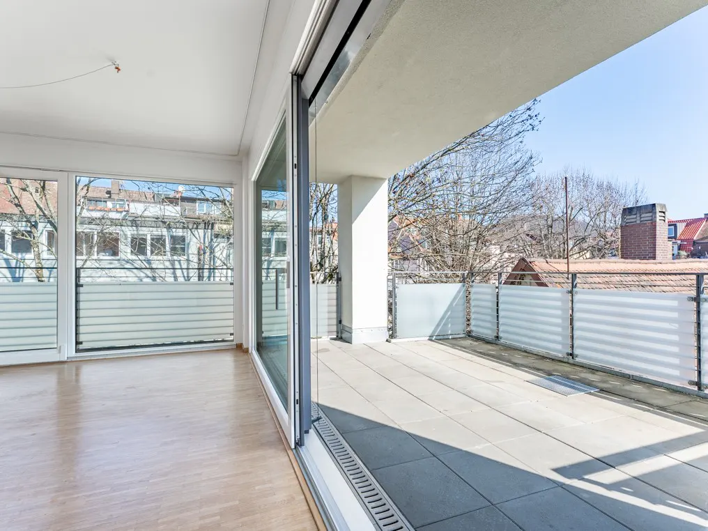 Bright, empty room with wood floors and sliding glass doors to a balcony. Balcony has gray tile and a frosted glass railing with a view of rooftops.