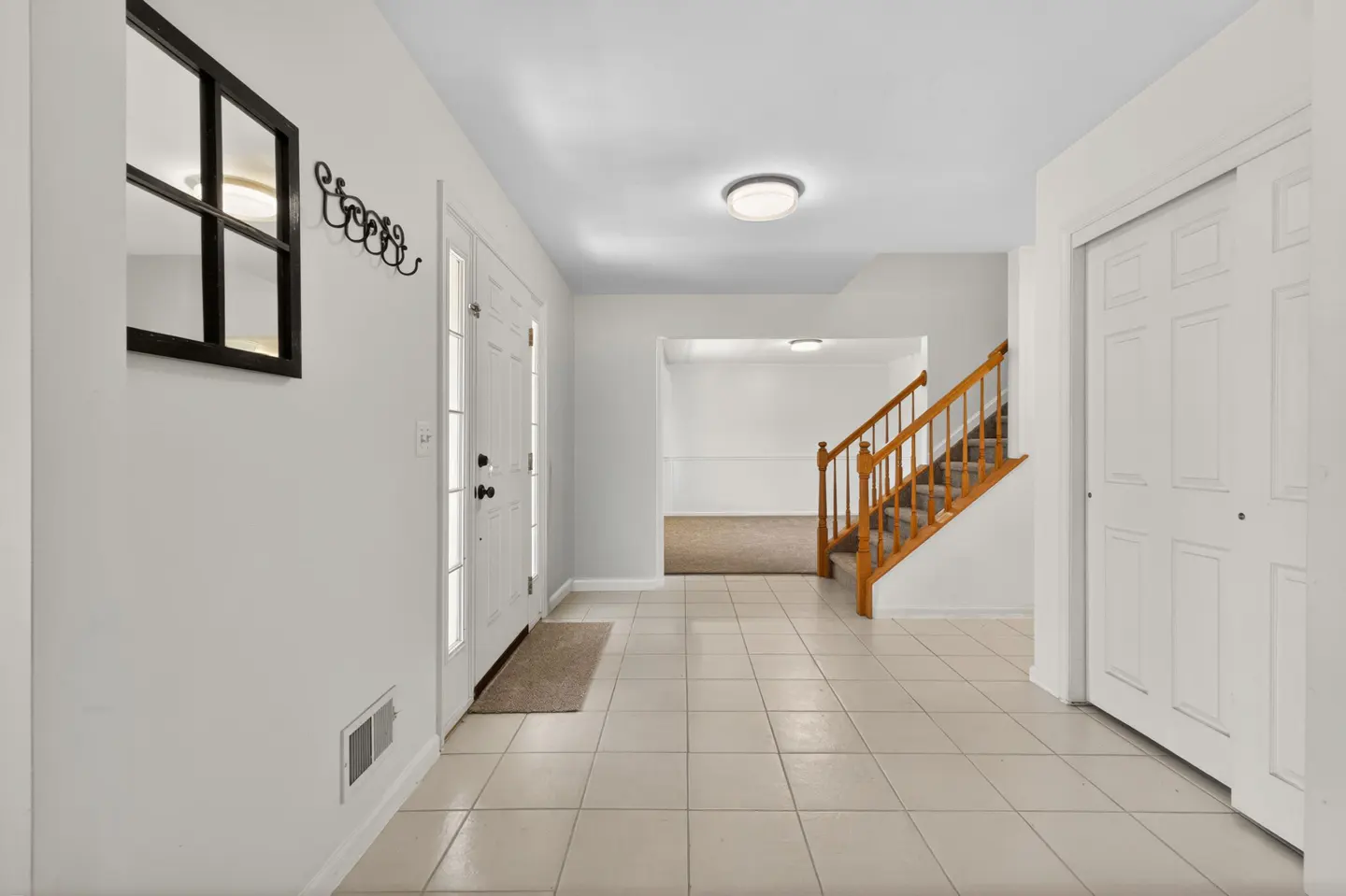 Bright entryway with tiled floor, white front door, wooden staircase, and a wall-mounted mirror and coat rack.