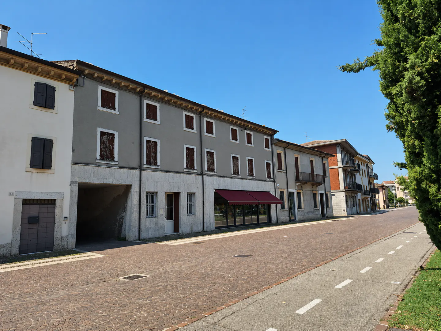 Street view of multi-story buildings with closed shutters, a red awning, and a brick road under a clear blue sky.