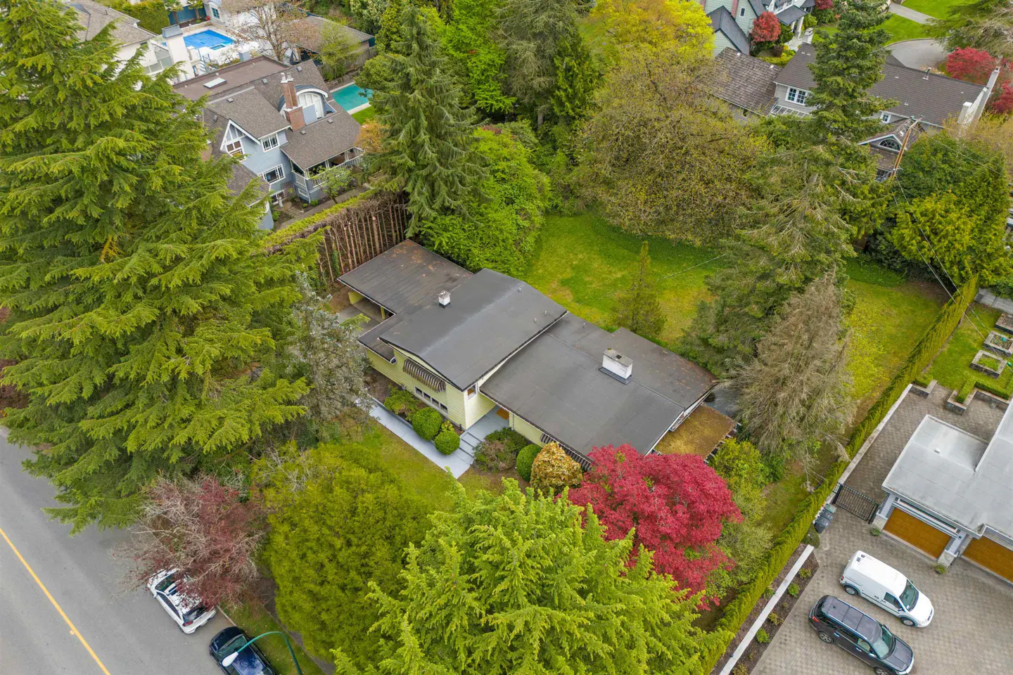 Aerial view of a yellow house with a dark roof, surrounded by green trees and a lawn. Cars are parked on the street.