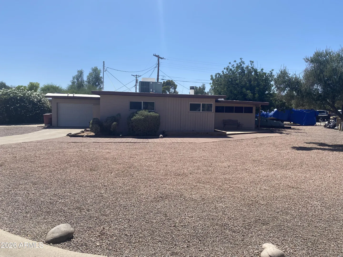 Single-story tan house with a flat roof, attached garage, and a large gravel front yard under a clear blue sky.