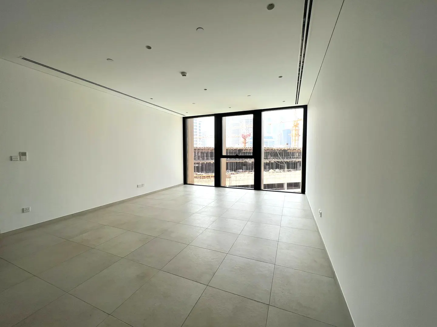 Empty, modern room with white walls, light tile floors, and large windows overlooking a city construction site.