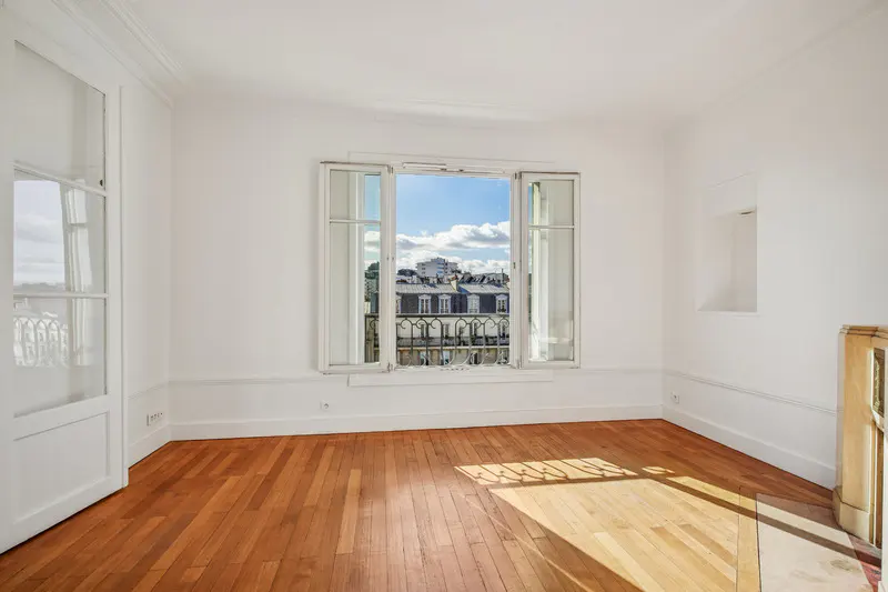 Bright, empty room with hardwood floors and white walls. Open window shows a city view. Sunlight shines on the floor.