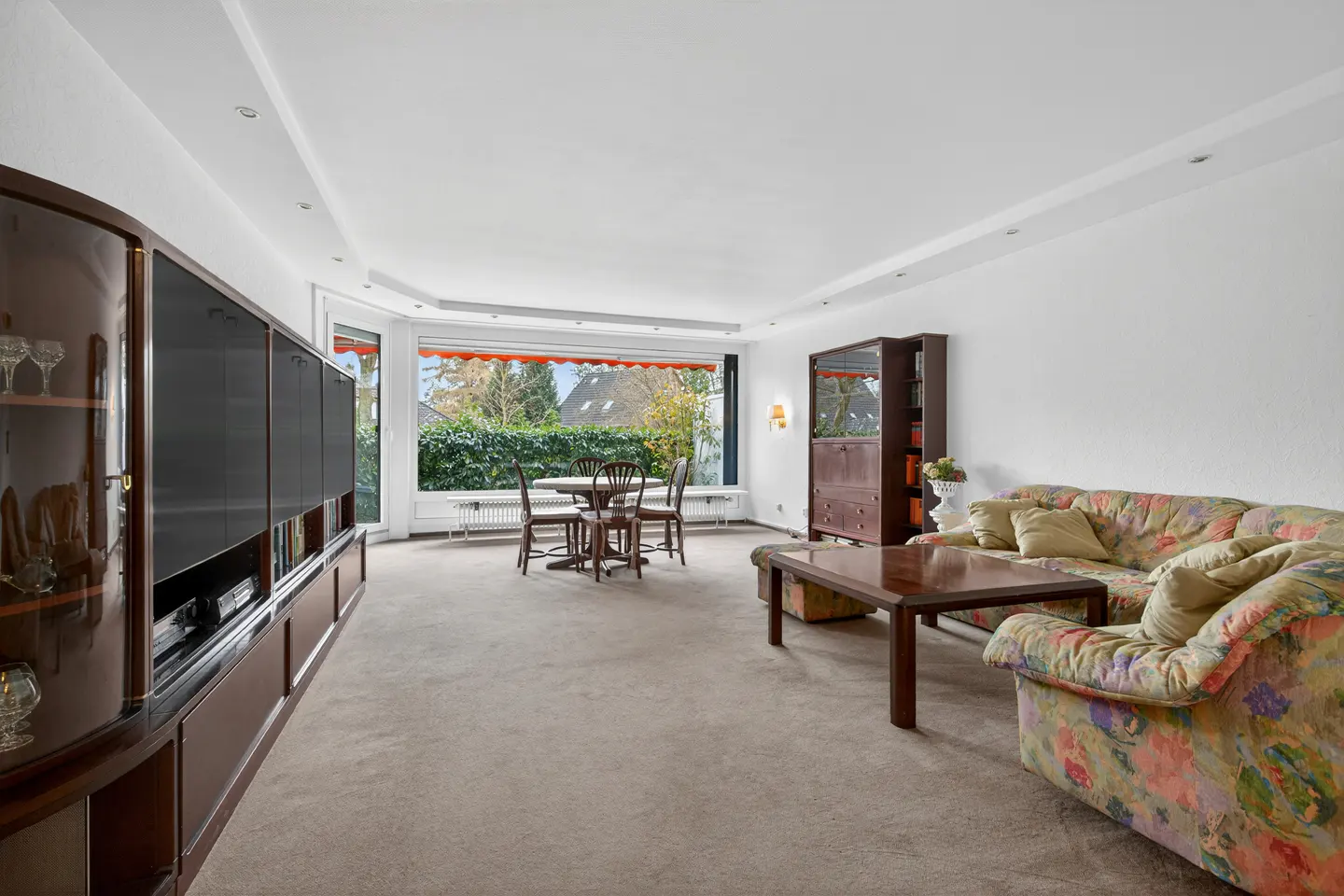 Bright living room with floral sofa, wood table, and built-in cabinet. A dining set sits near a large window with an orange awning.