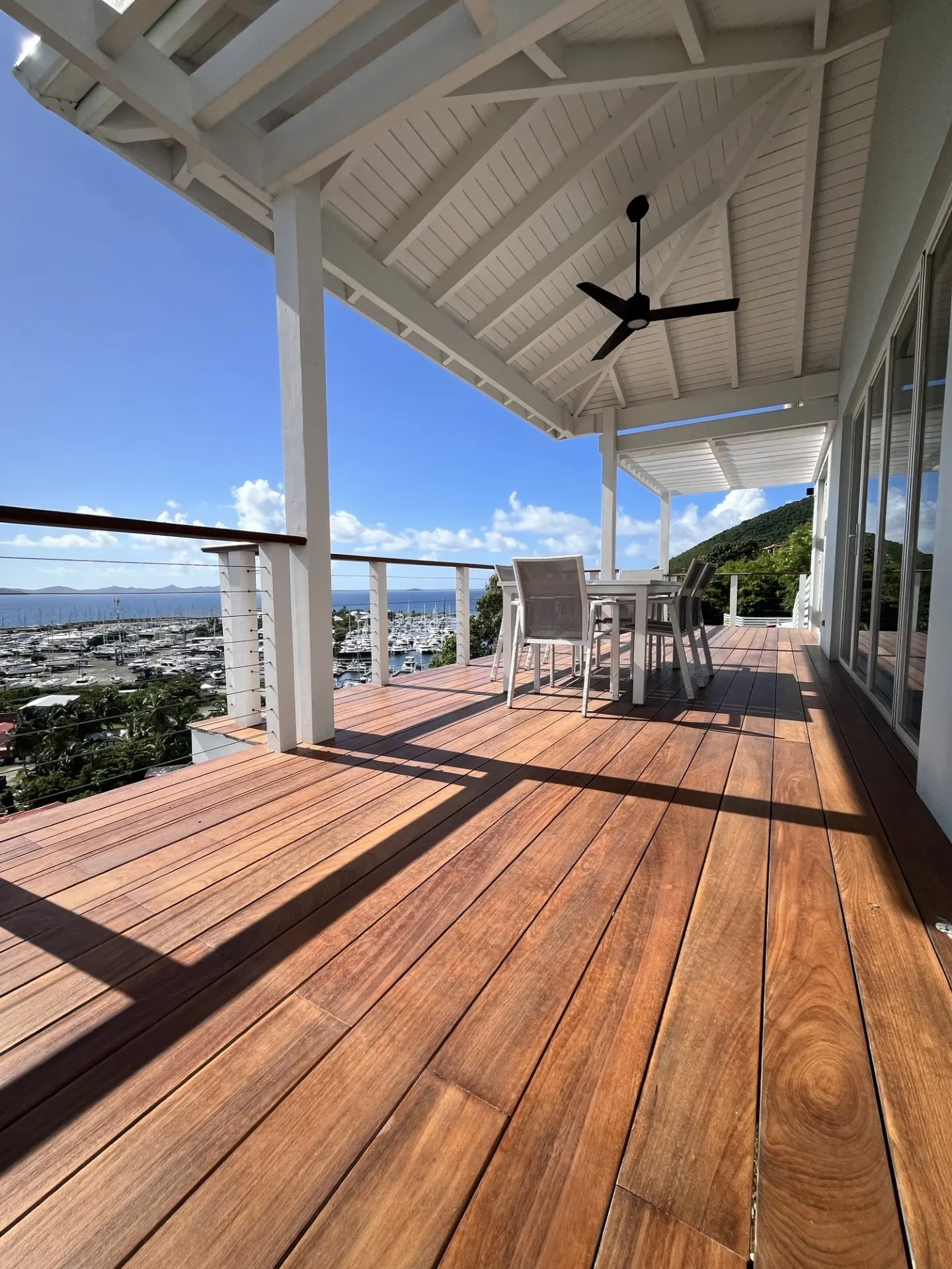 Wide angle view of a wood deck with white ceiling, pillars, and a black ceiling fan overlooking a marina and blue sky.