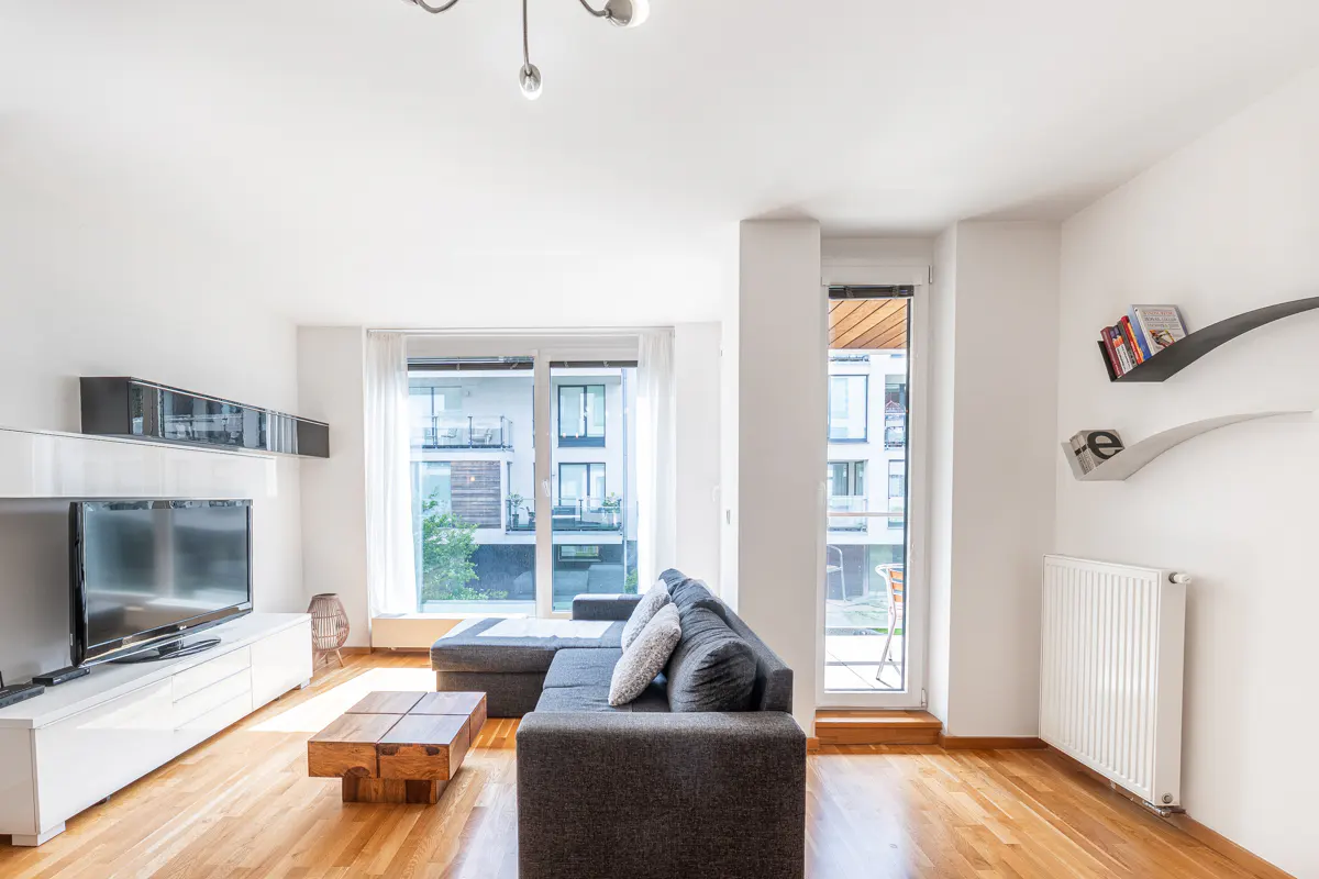 Bright living room with gray sectional sofa, wood floors, and a large window with a view of an apartment building.