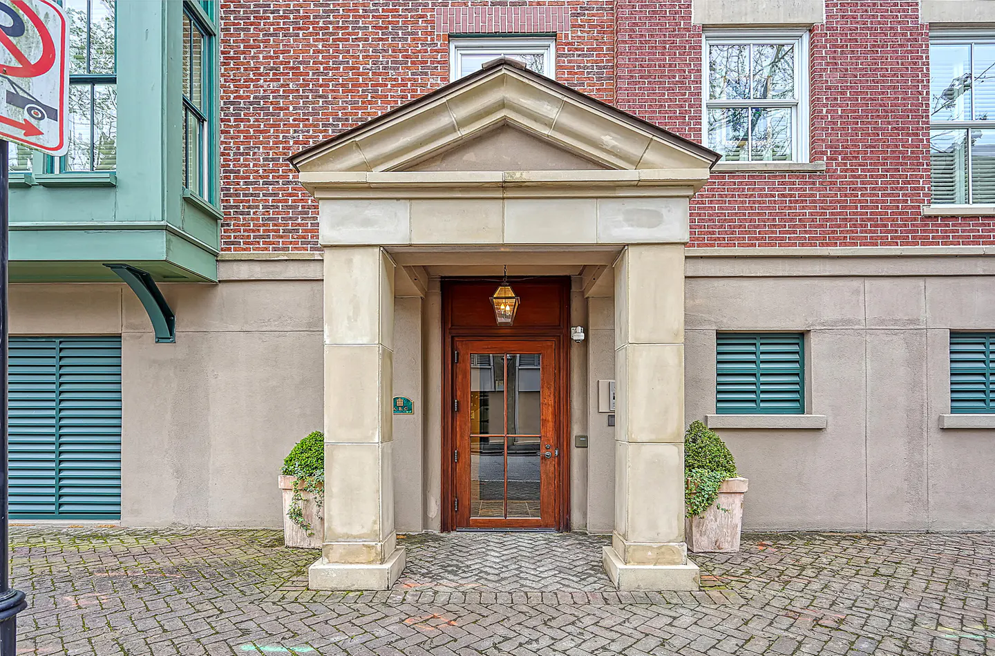 Building entrance with a brown door framed by stone pillars and a hanging lantern. Red brick facade and green shutters.