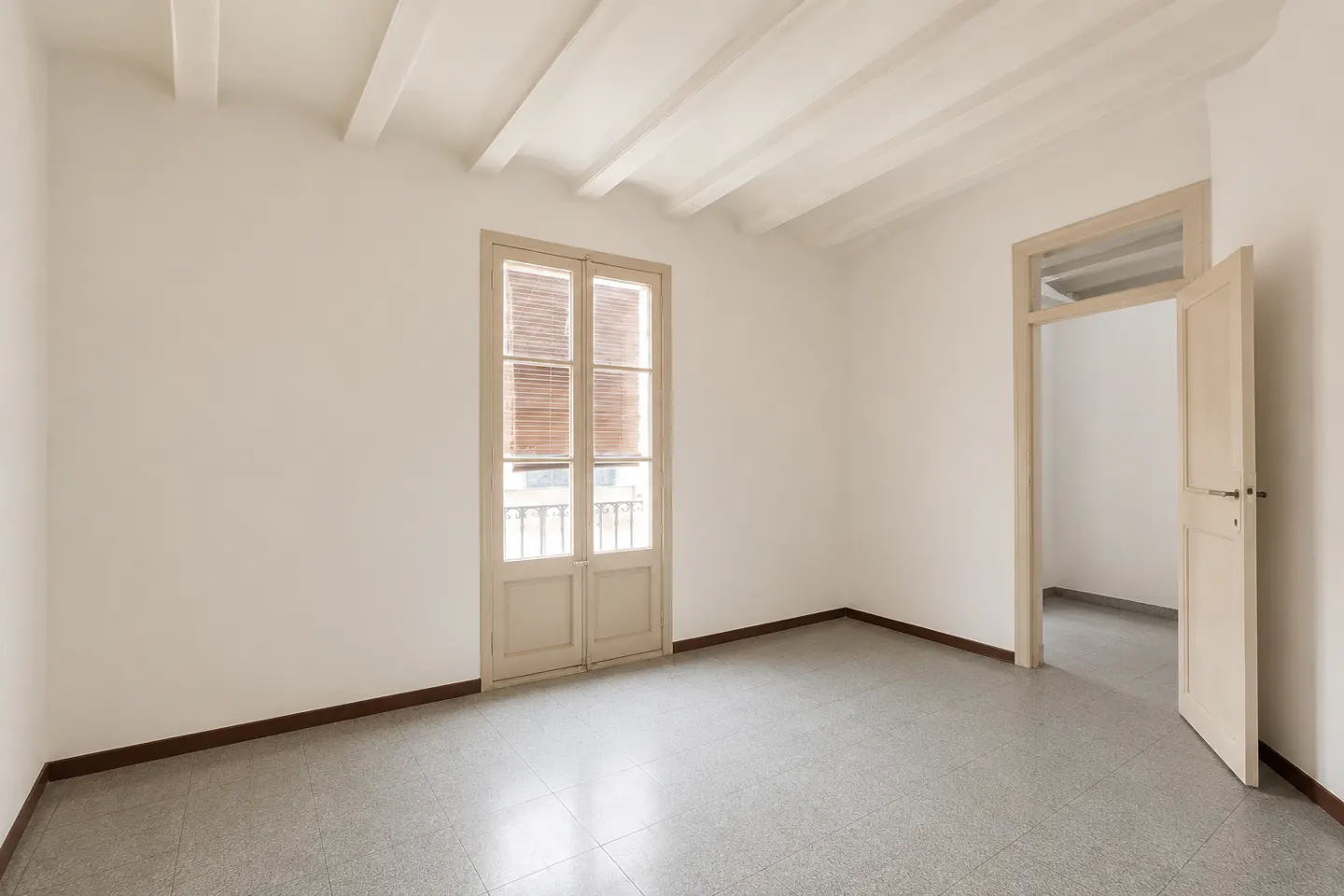 Empty room with white walls, exposed ceiling beams, and gray tile floor. A double door with blinds leads to a balcony. An open door leads to another room.