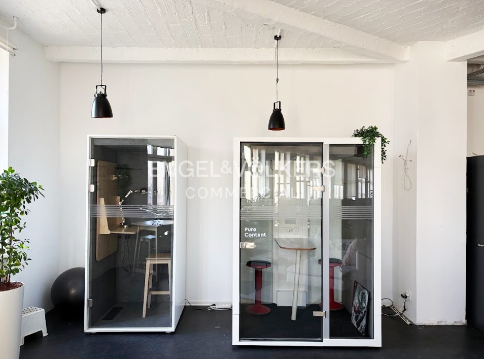 Two modern glass office phone booths with stools and desks sit against a white wall under black pendant lights.