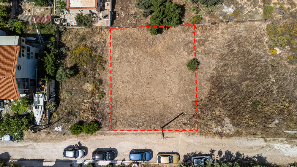 Aerial view of a rectangular vacant plot of land outlined in red, situated next to a house and a street with parked cars.