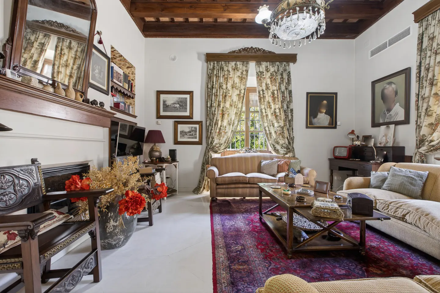 A living room with floral curtains, a wooden ceiling, and a red rug. There are sofas, a coffee table, and a fireplace with a mirror above it.