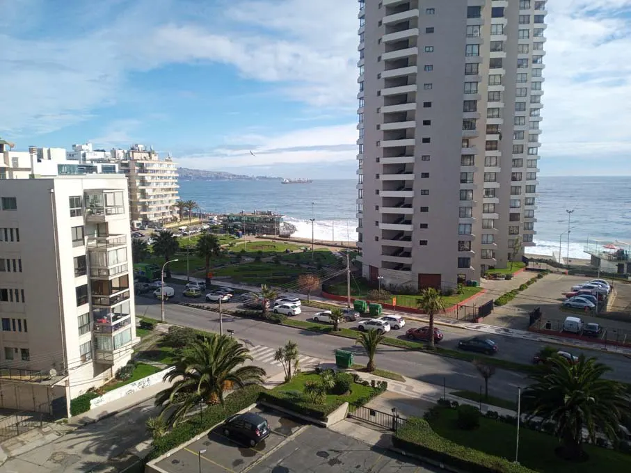 View of a coastal city with tall buildings, a park, and the ocean under a blue sky.