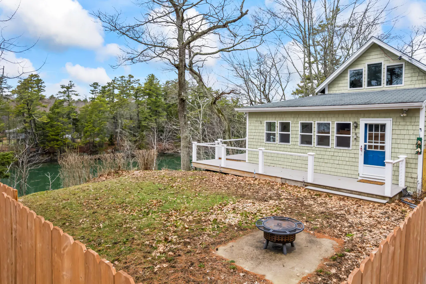 Exterior view of a light green house with a blue door, a wooden deck, and a fire pit in the yard.