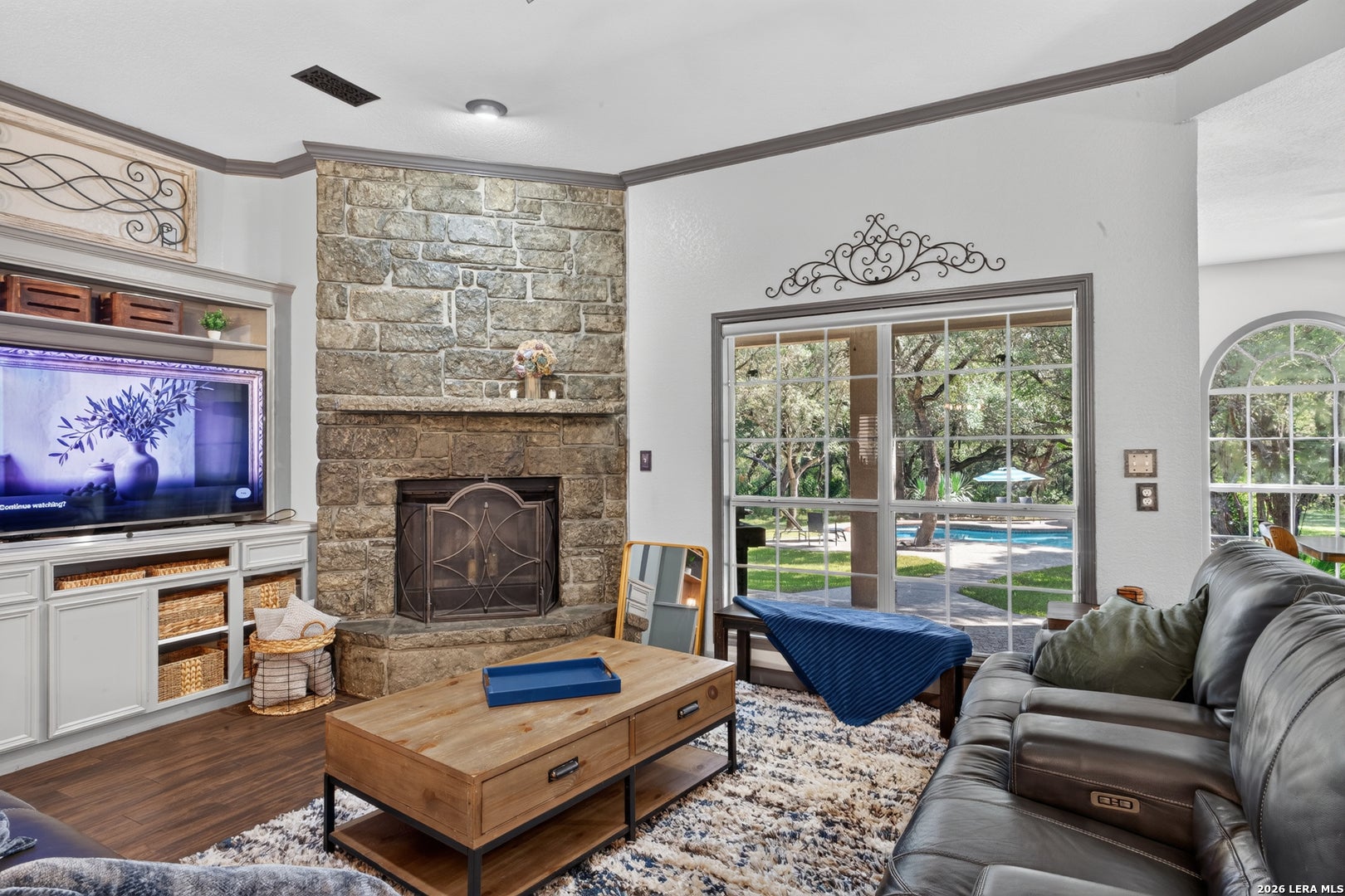 Living room with stone fireplace, TV console, wooden coffee table, leather sofa, and large windows overlooking a pool. [AI-generated text]