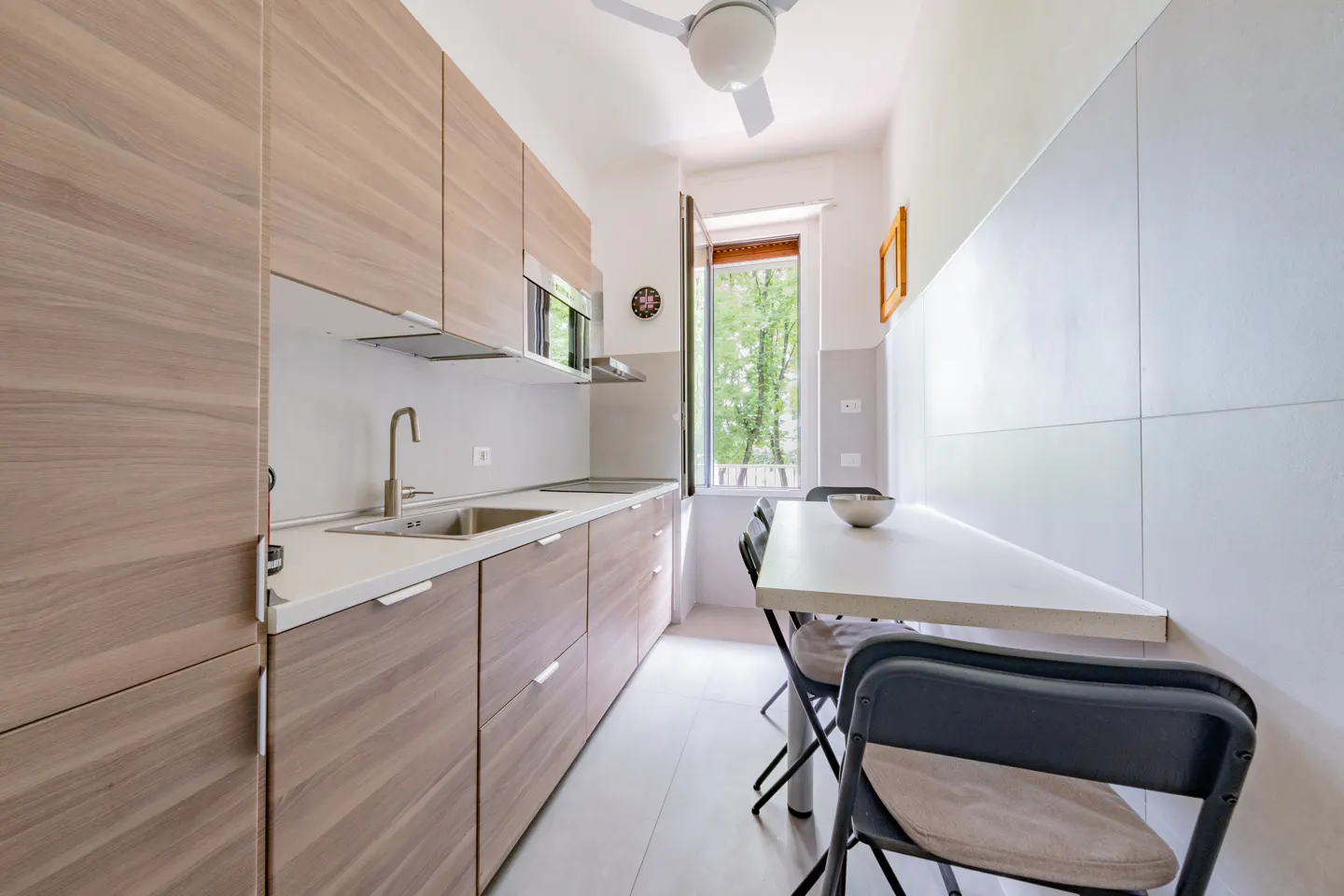 Modern galley kitchen with light wood cabinets, white countertops, a small dining table, and a window with a green view.