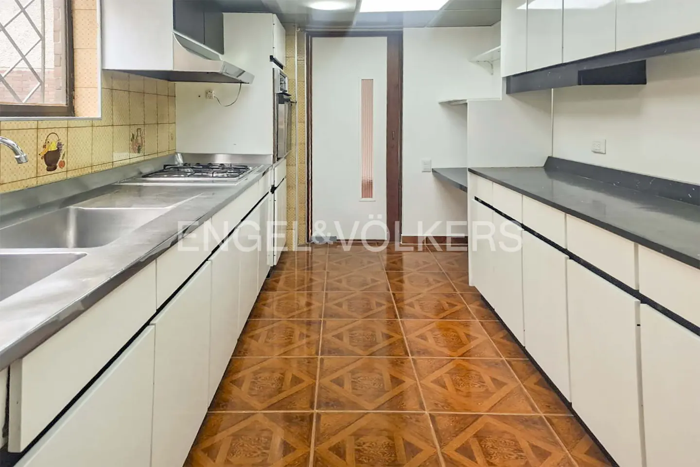 Galley kitchen with white cabinets, dark countertops, stainless steel sink, gas stove, and brown patterned tile flooring.