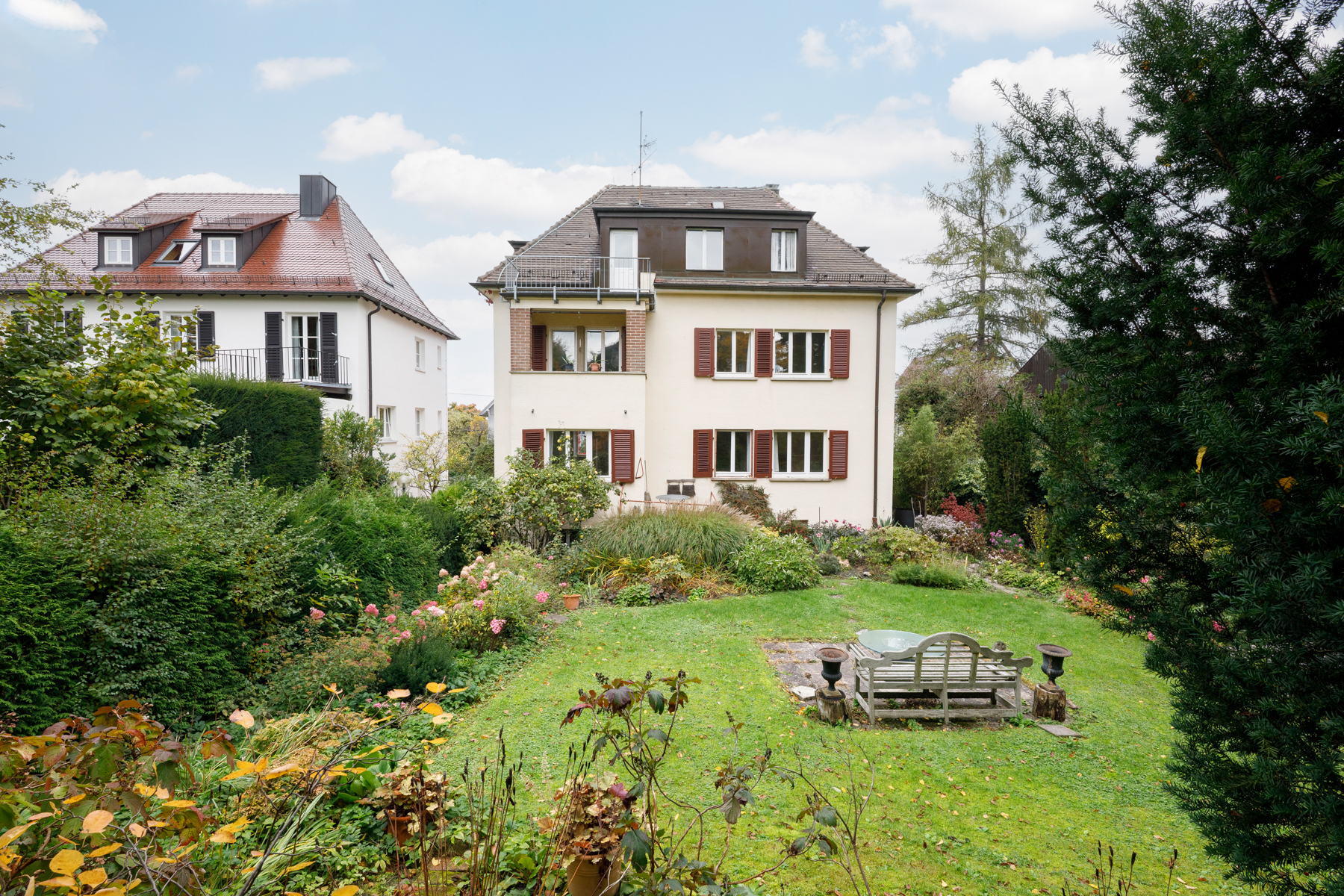 A two-story cream house with brown shutters and a balcony, surrounded by a green lawn and lush garden. A wooden bench sits on the grass.