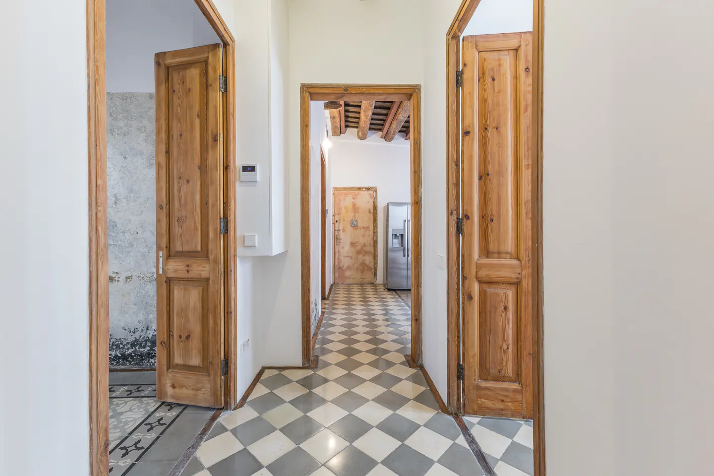 Hallway with checkered tile floor, white walls, and three open wood doors. A stainless steel refrigerator is visible in the background.