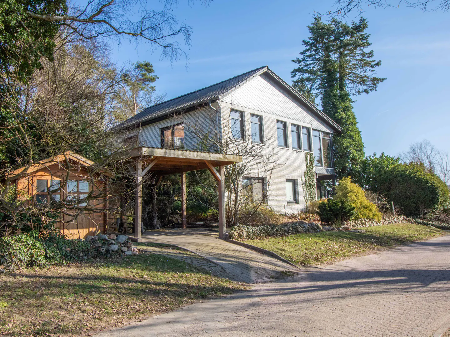Two-story house with gray brick, dark roof, and carport. A small wooden shed sits to the left, surrounded by trees and greenery.