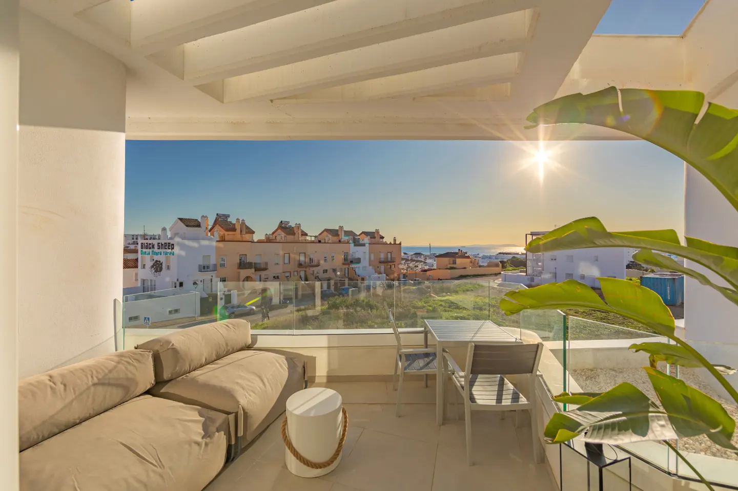 Balcony view with beige sofa, white stool, and table set. Ocean and buildings visible in the background under a sunny sky.