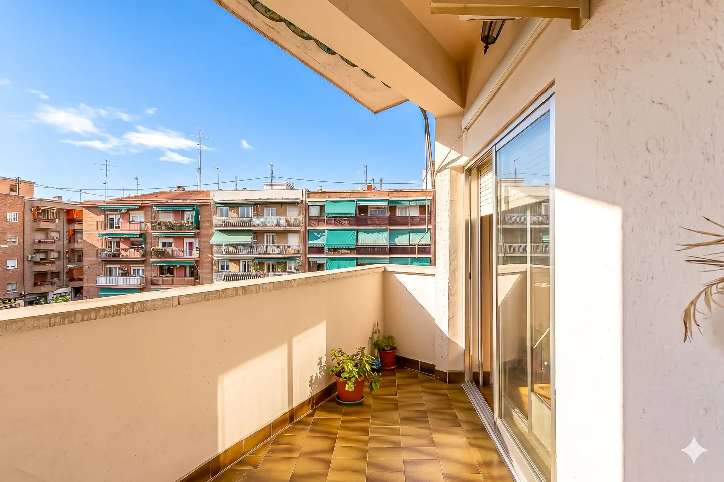 Balcony view with brown tiled floor, white walls, potted plants, and sliding glass doors. Buildings visible in the background under a blue sky.