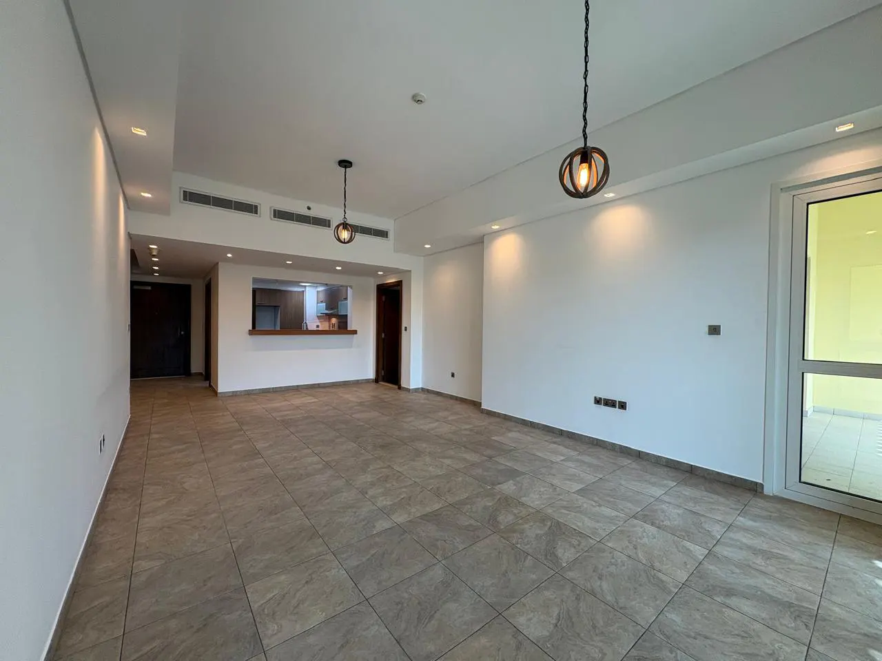 Bright, empty living room with tile floors, white walls, and hanging pendant lights. A kitchen is visible through a serving hatch.