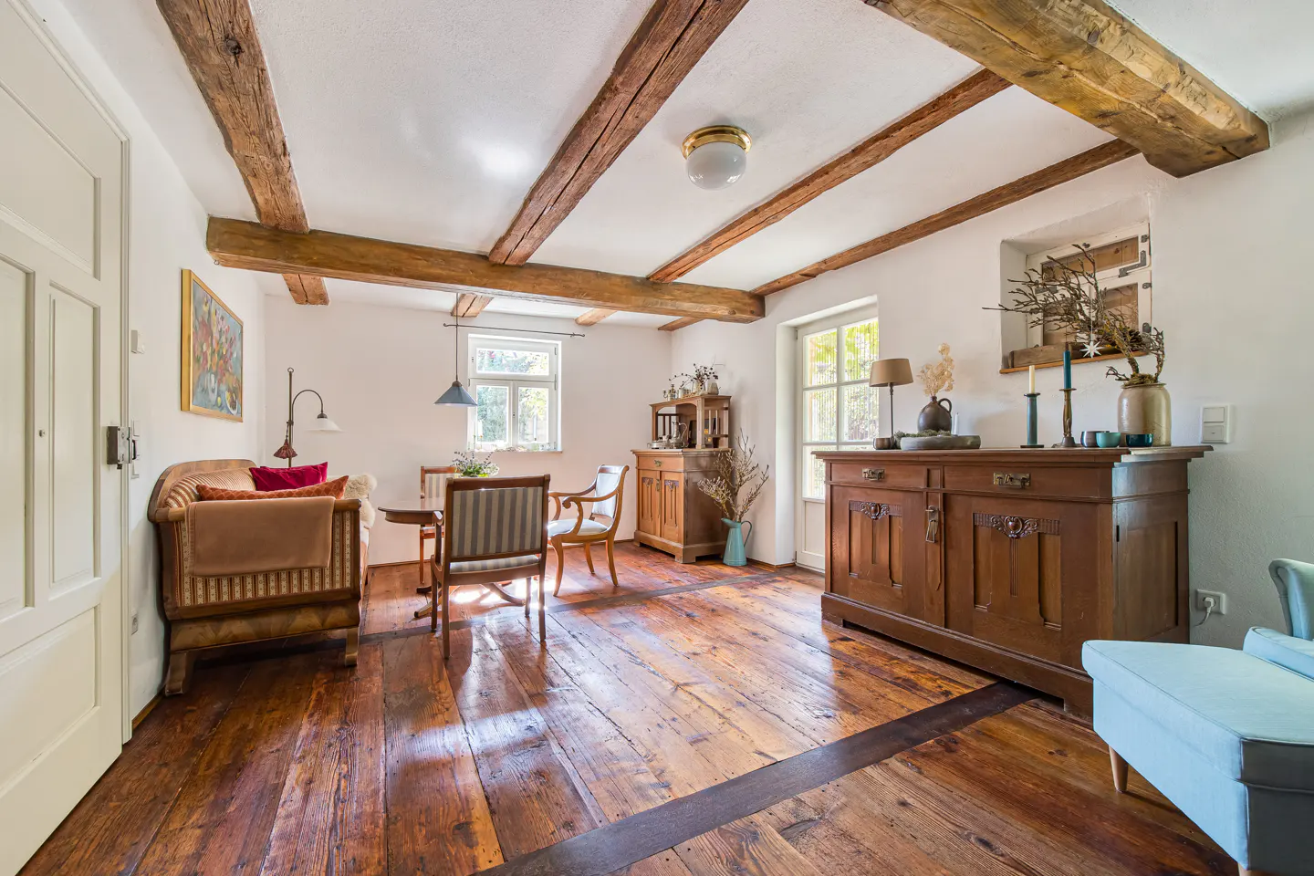 Living room with exposed wood beams, hardwood floors, and antique furniture. Windows provide natural light.