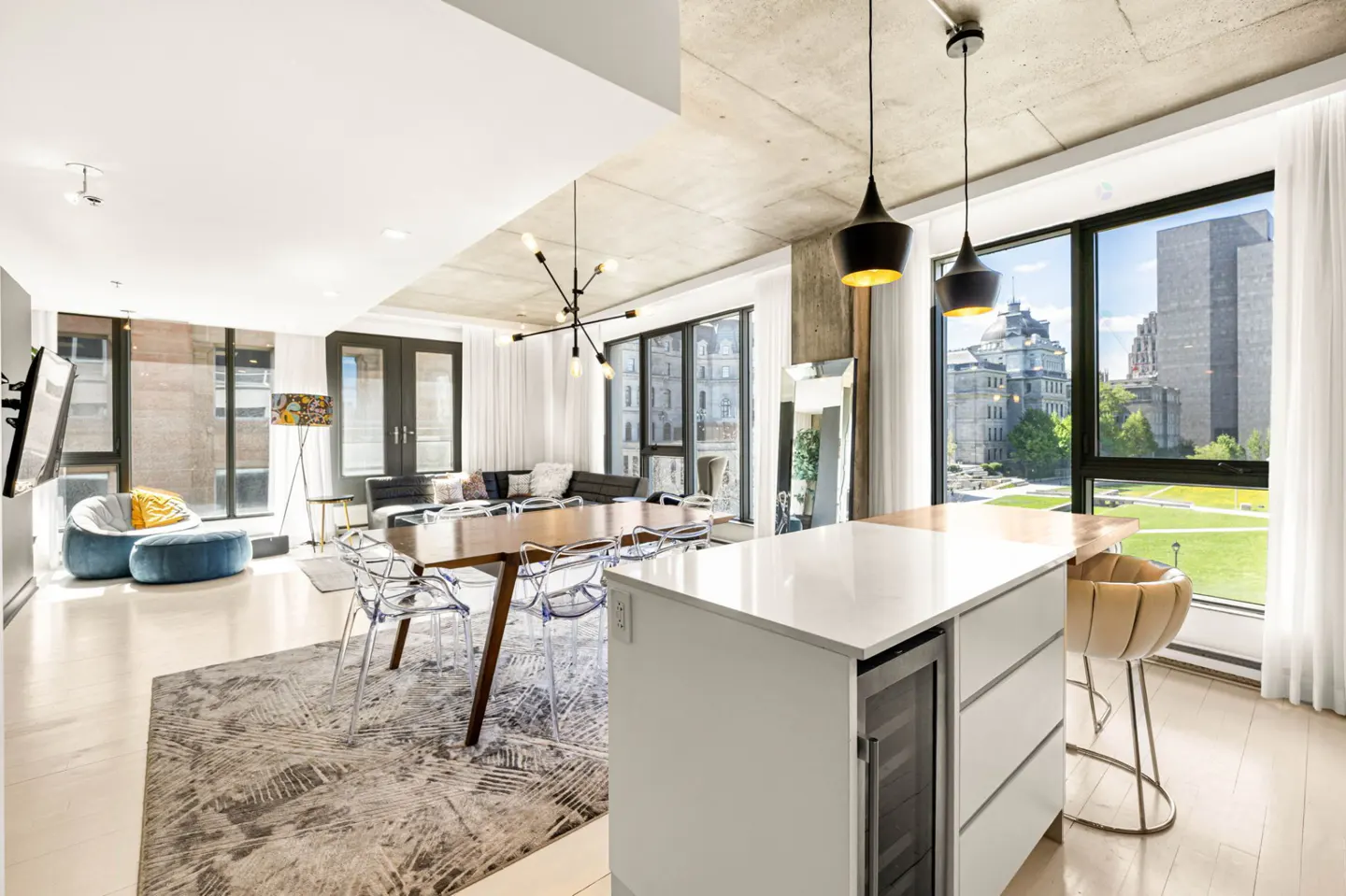 Open-concept living space with a dining table, kitchen island, and seating area. Large windows offer city views. Neutral color palette.