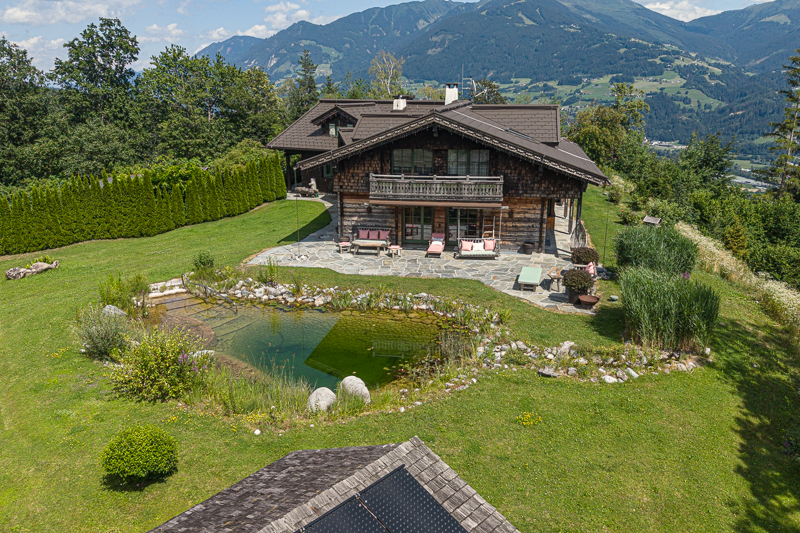 A rustic wooden chalet with a stone patio, natural swimming pond, and lush lawn, set against a backdrop of alpine mountains.