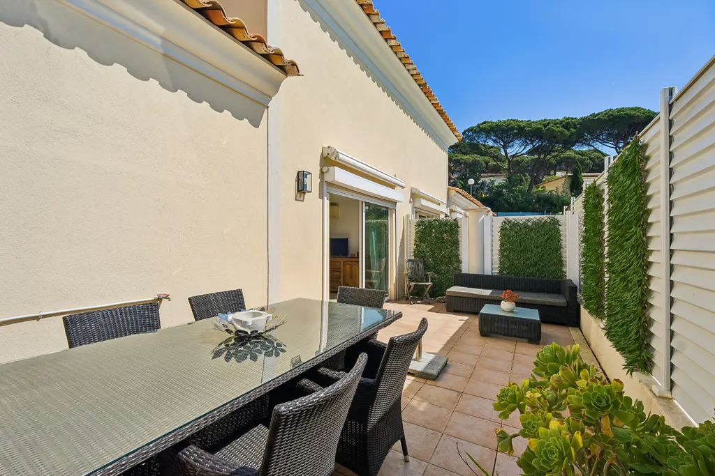 Outdoor patio with a glass table, wicker chairs, and a black sofa set against a cream-colored building and a blue sky.