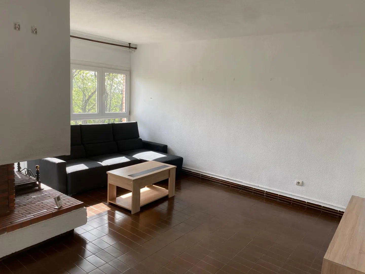 Bright living room with dark wood floors, white walls, and a black sectional sofa. A light wood coffee table sits in front of the sofa. A brick fireplace is on the left.