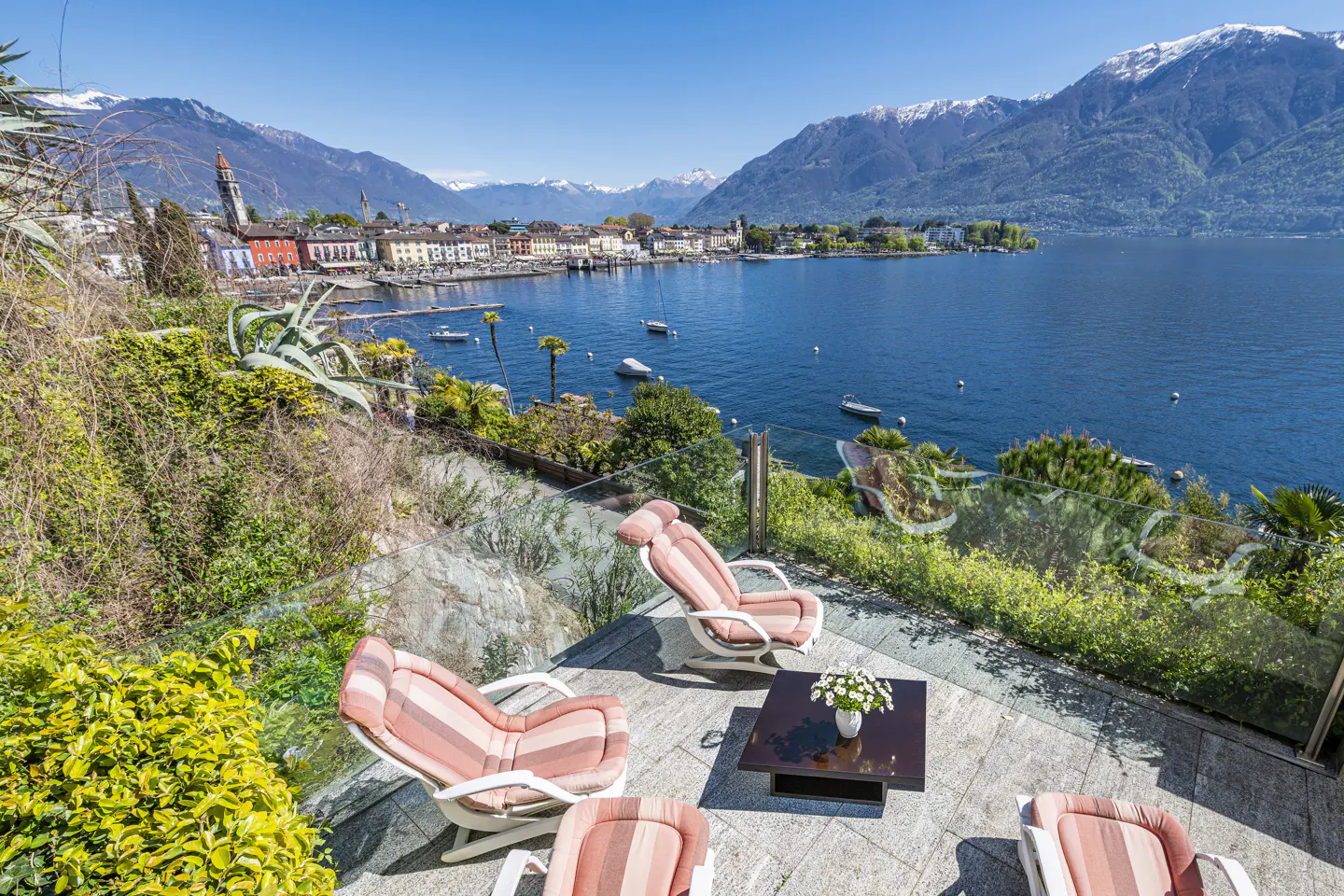 Patio with striped lounge chairs overlooking a blue lake, town, and snow-capped mountains on a sunny day.