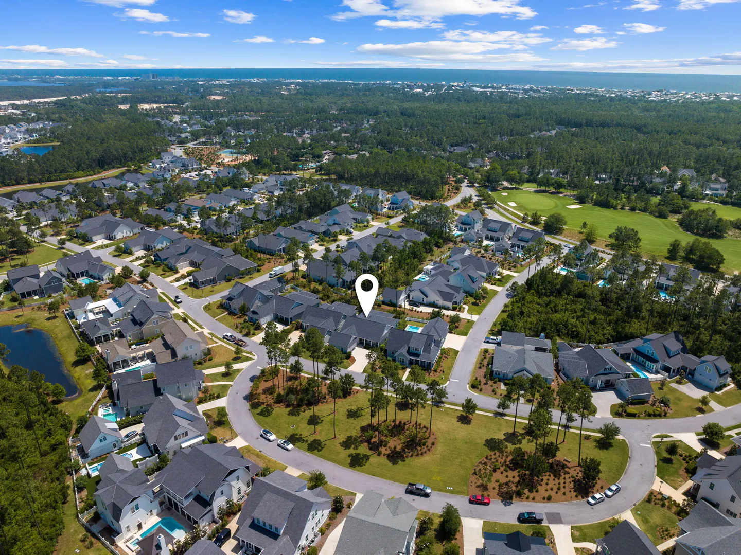 Aerial view of a residential neighborhood with gray roofed houses, green trees, and a golf course in the background. A location pin is centered.