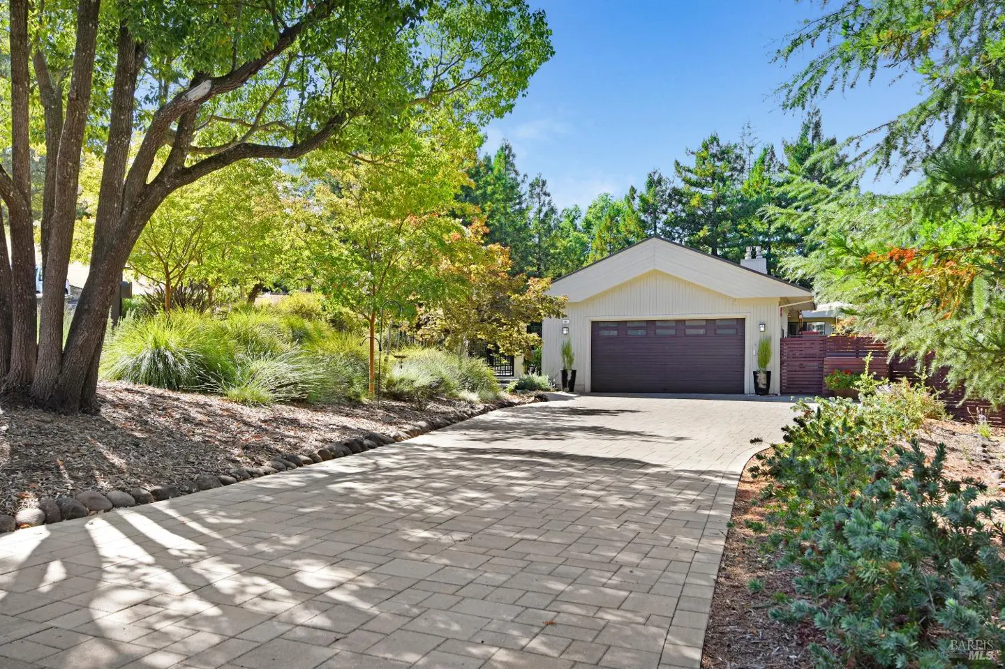 A single-story house with a brown garage door and a brick driveway, surrounded by green trees and plants.