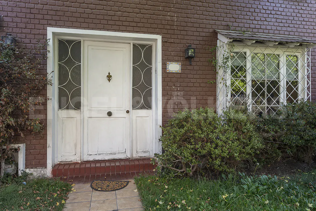 Exterior shot of a brick house with a white front door and diamond-patterned windows. A small, round doormat sits on the stone walkway leading to the entrance.
